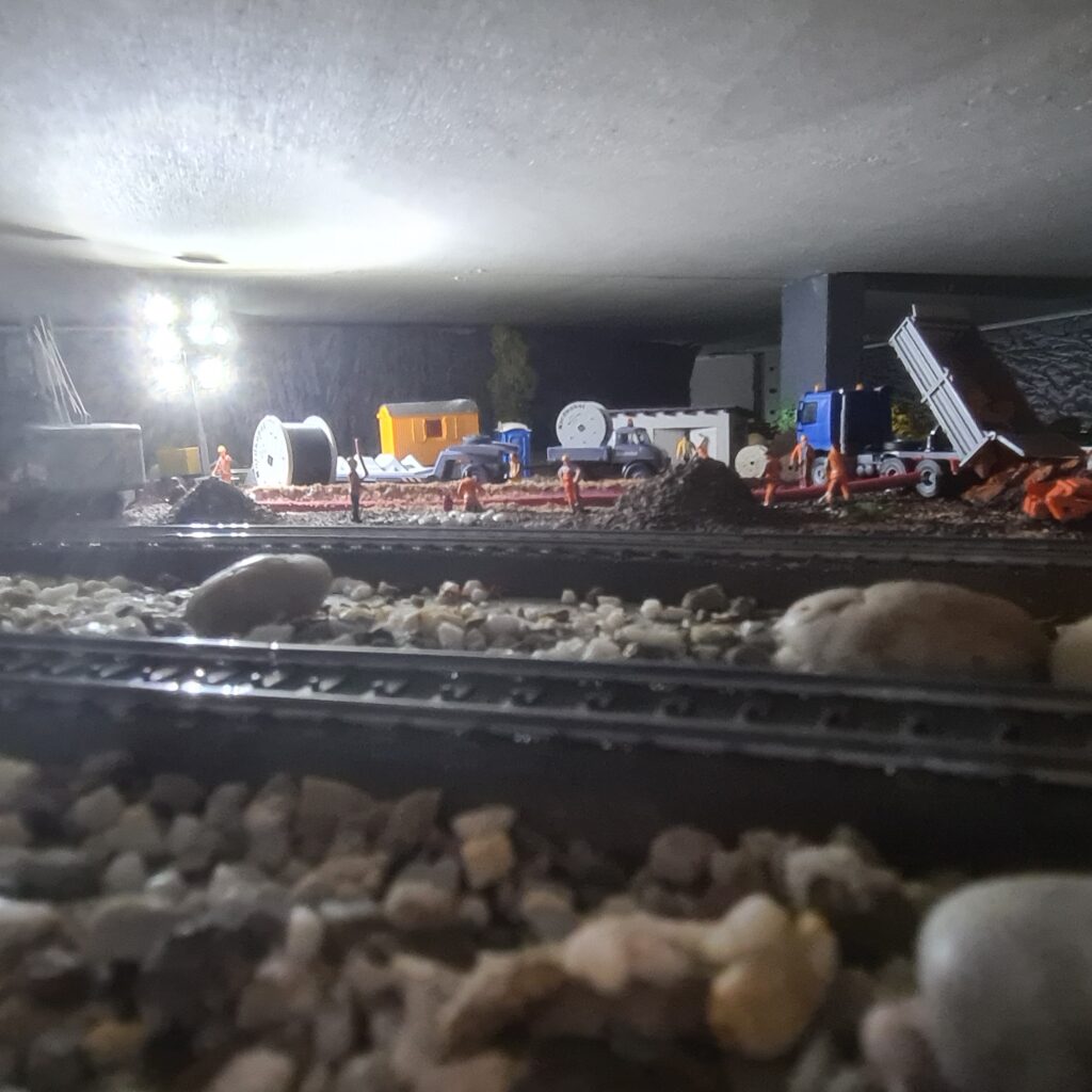 Blick in einen Modellbahntunnel auf eine beleuchtete Baustellenszene mit Baufahrzeugen und Figuren.