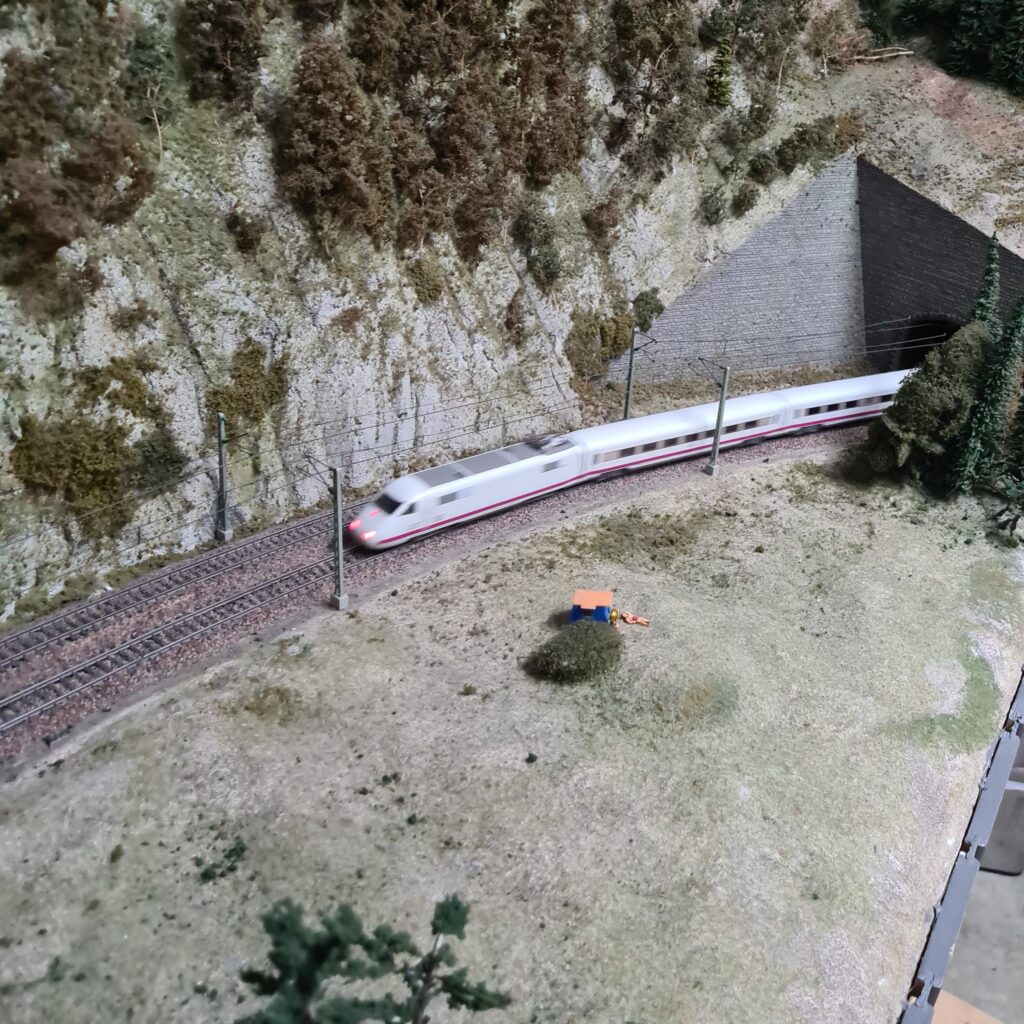Weißer Hochgeschwindigkeitszug fährt entlang einer Felswand aus einem Tunnel.