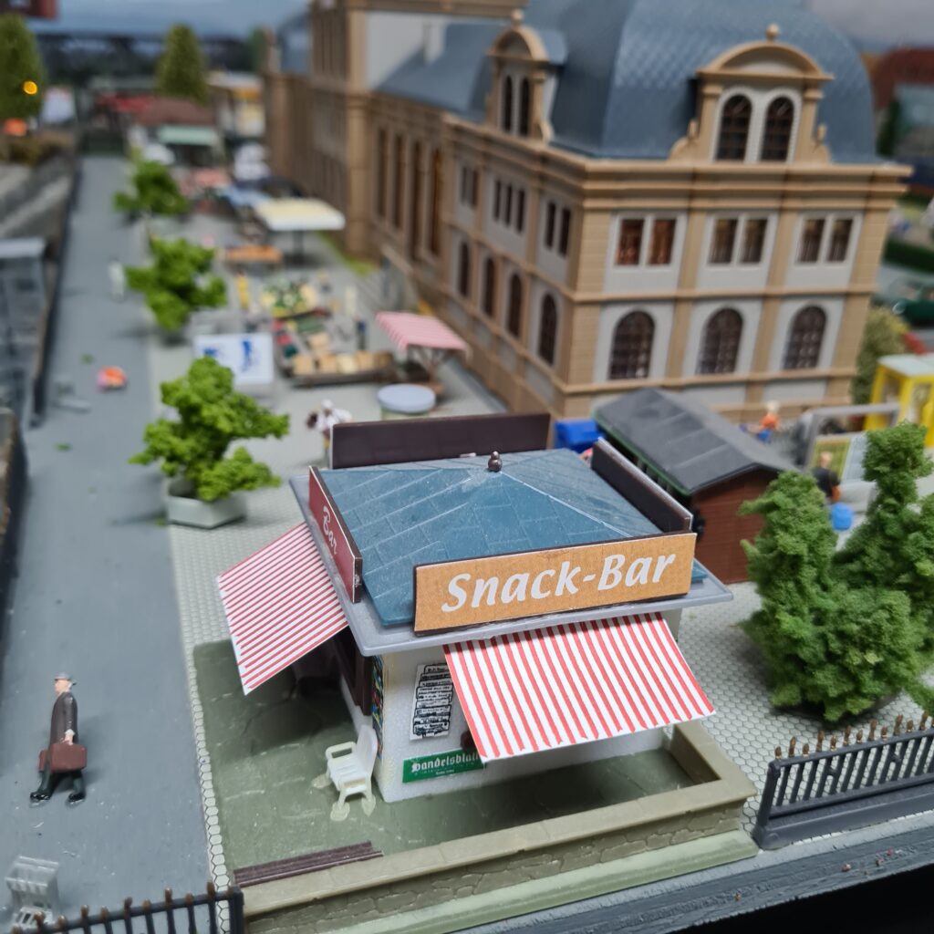 Snack-Bar mit rot-weiß gestreiften Markisen vor dem Bahnhofsgebäude der Modellbahnanlage.