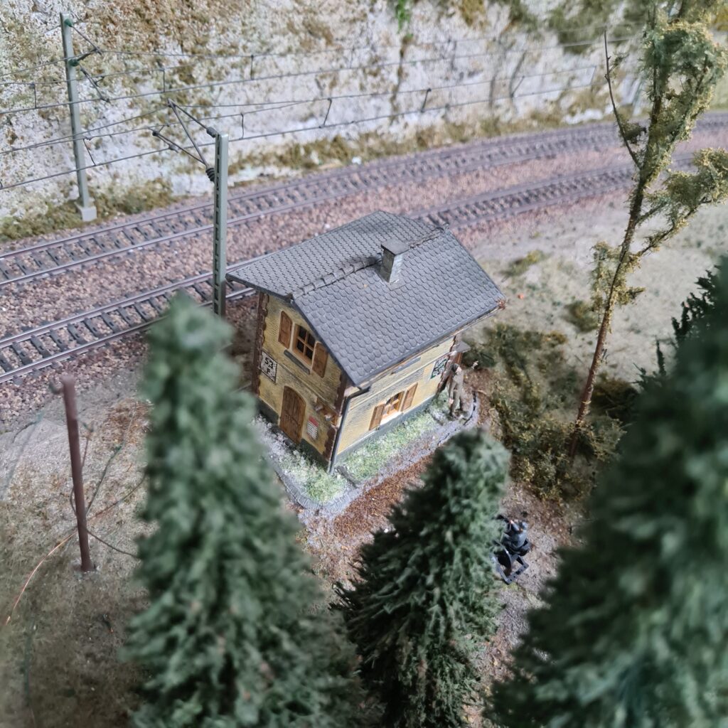 Kleines Haus zwischen Bäumen direkt neben einer elektrifizierten Bahnstrecke.