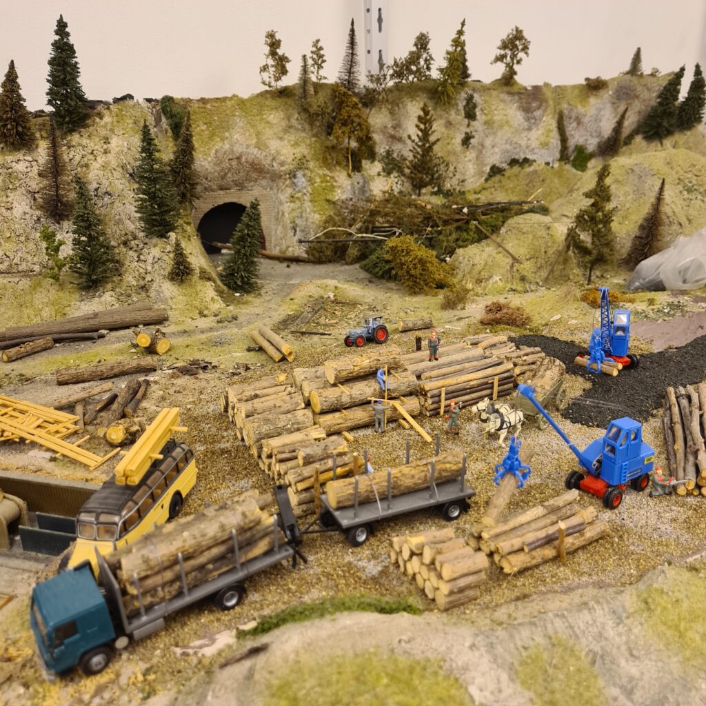 Holzverladung mit Baumstämmen, Lastwagen und Arbeitsfahrzeugen vor einer bewaldeten Felslandschaft.