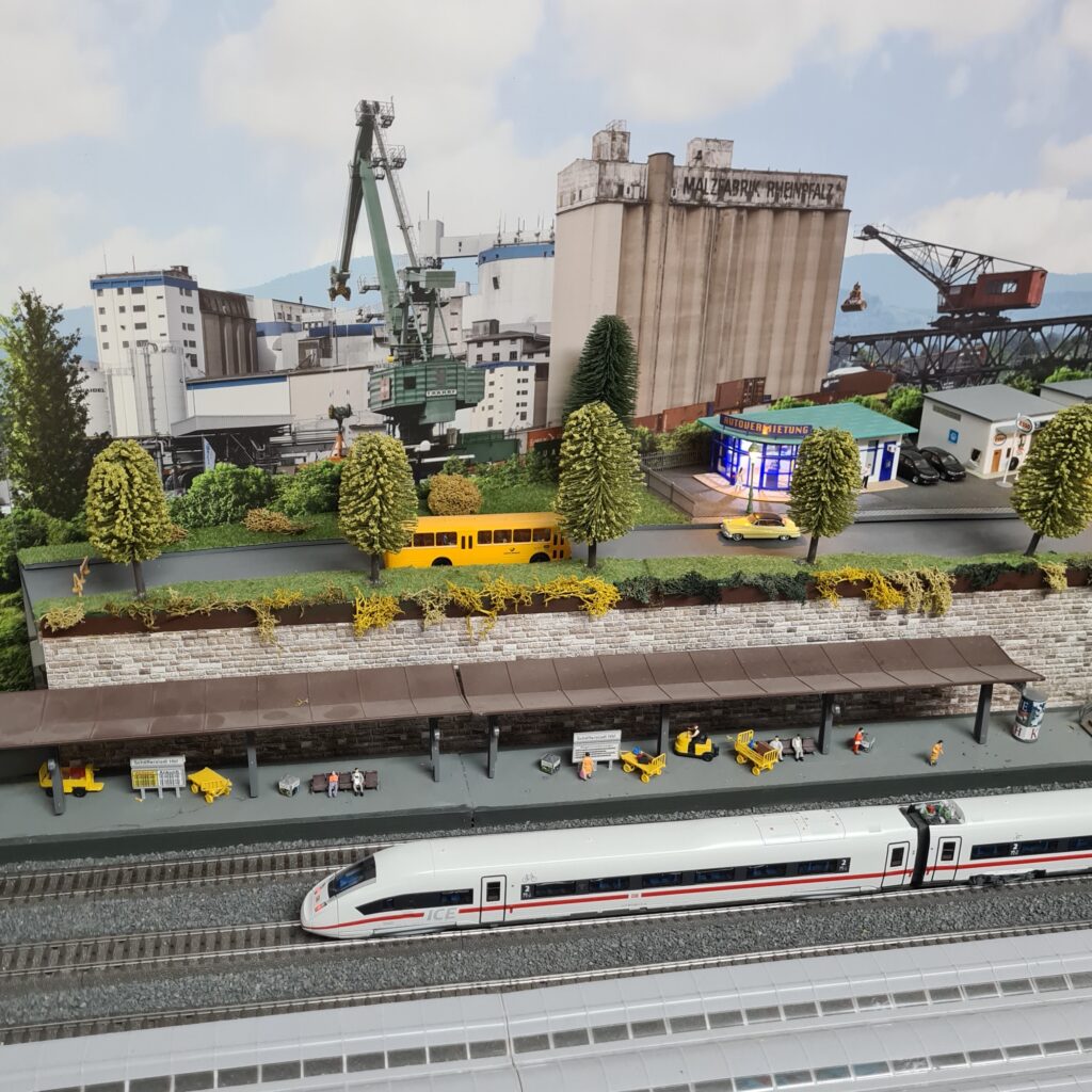 ICE am Bahnsteig der Modellbahnanlage, dahinter eine Industrieanlage mit Silos und Kränen.