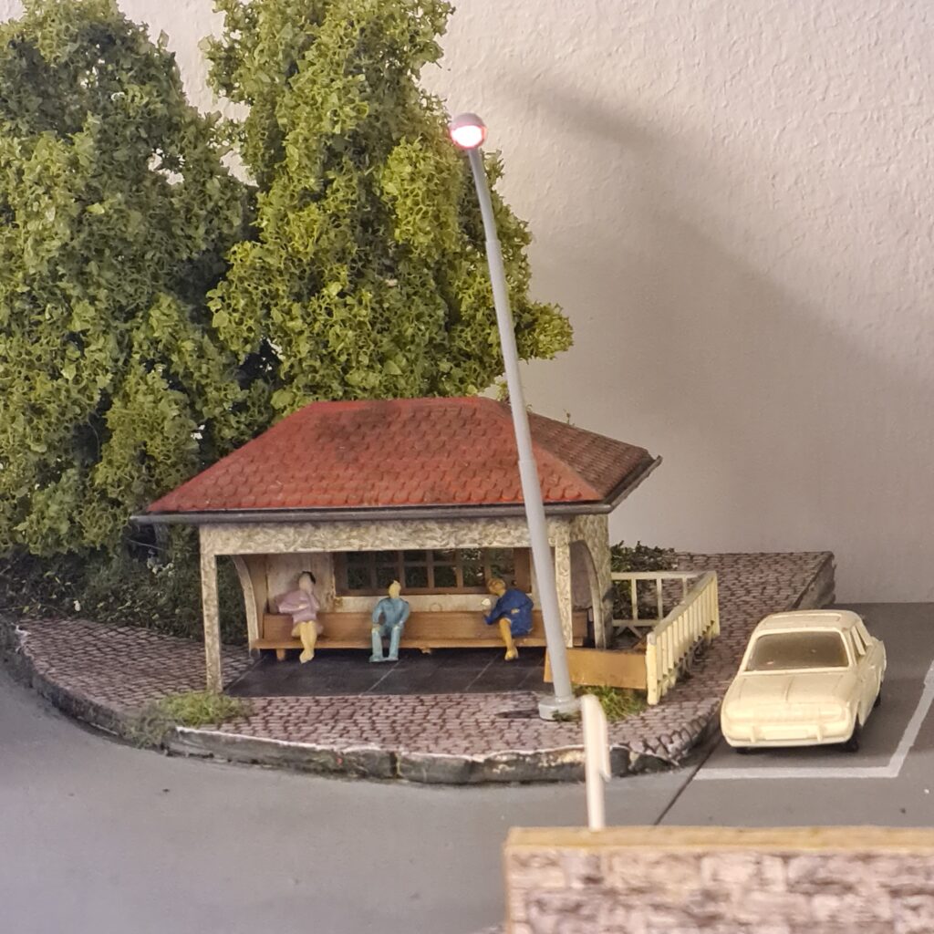 Überdachte Bushaltestelle mit wartenden Fahrgästen, Straßenlaterne und geparktem Auto.
