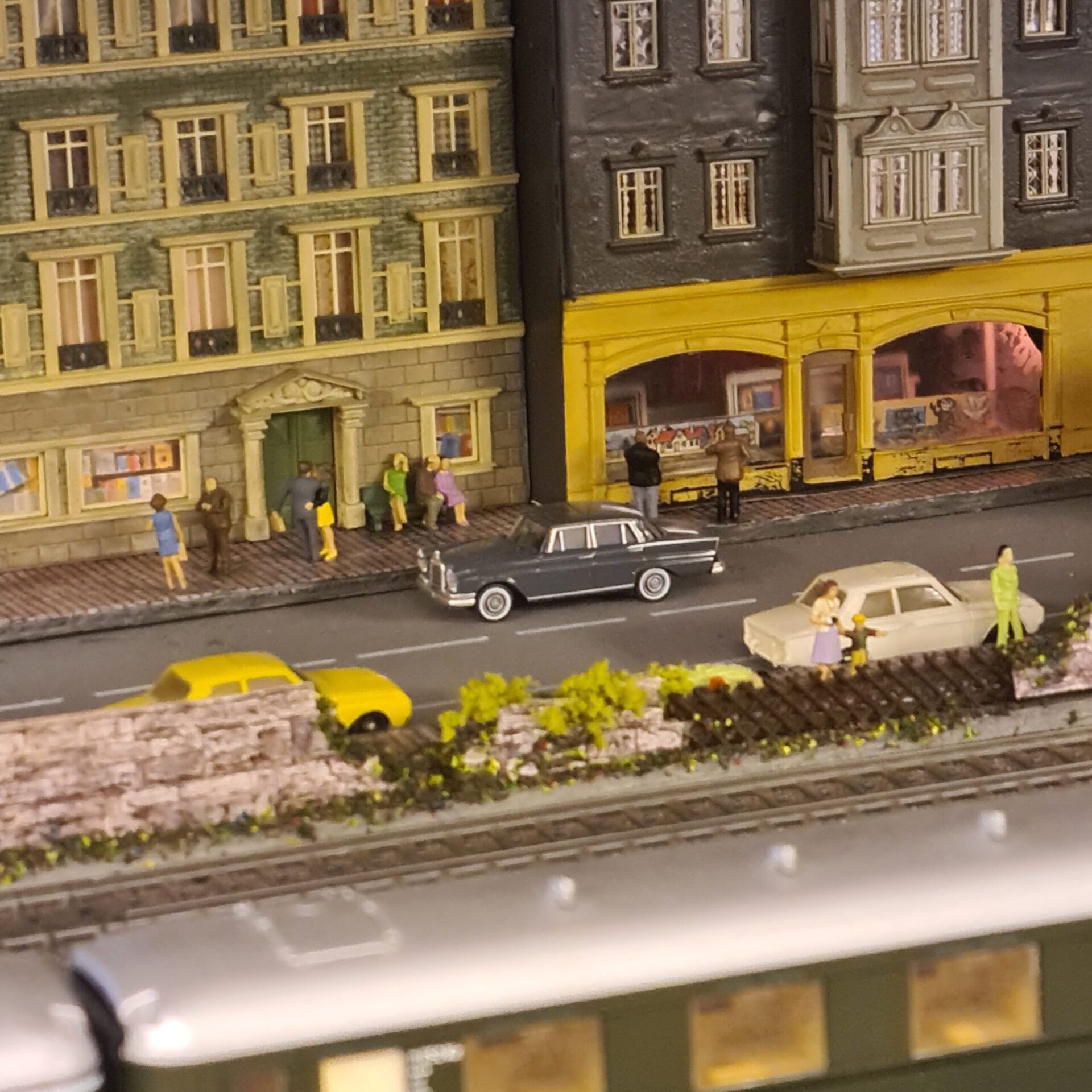 Straßenszene vor historischen Stadthäusern mit Geschäften, Passanten und parkenden Autos.