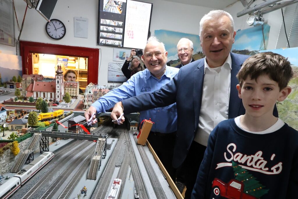 Bürgermeister Peter Burger und MEC-Vorsitzender Volker Raddatz stehen an der Modellbahnanlage und geben gemeinsam die Fahrstrecke frei. Weitere Gäste schauen zu.