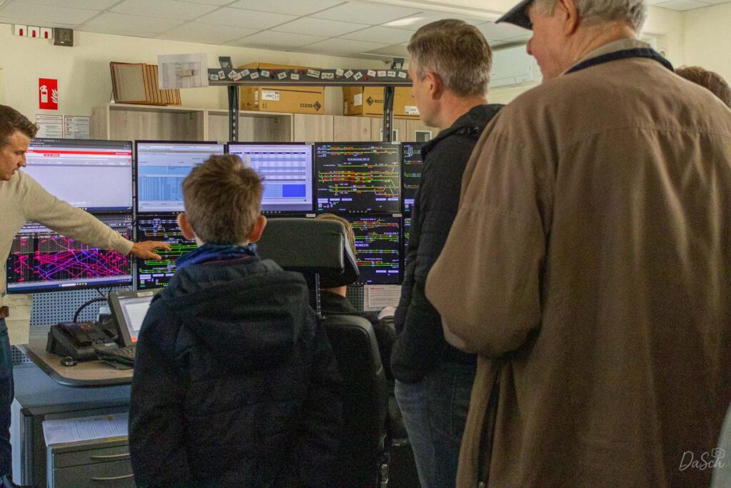 Besucher stehen vor mehreren Monitoren im Stellwerk der Riedbahn. Ein Mitarbeiter erläutert die angezeigten Gleis- und Steuerungsübersichten.