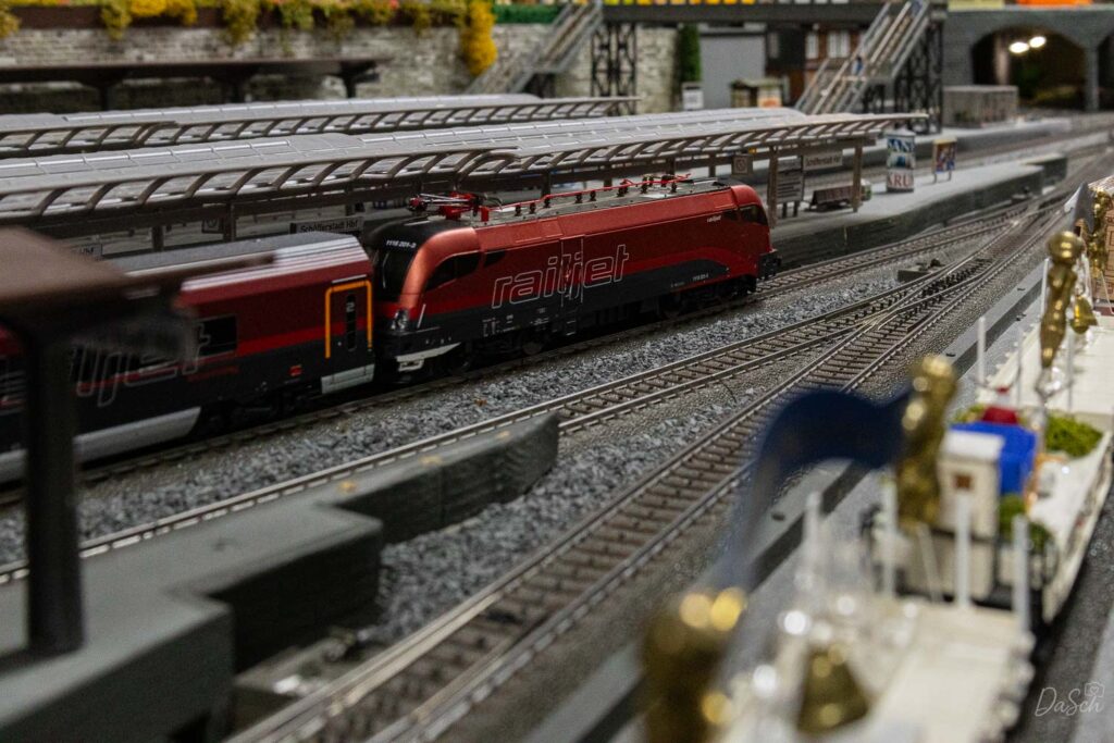 Rote Railjet-Modelllokomotive mit Reisezugwagen fährt auf den Gleisen des Modellbahnhofs an überdachten Bahnsteigen vorbei.