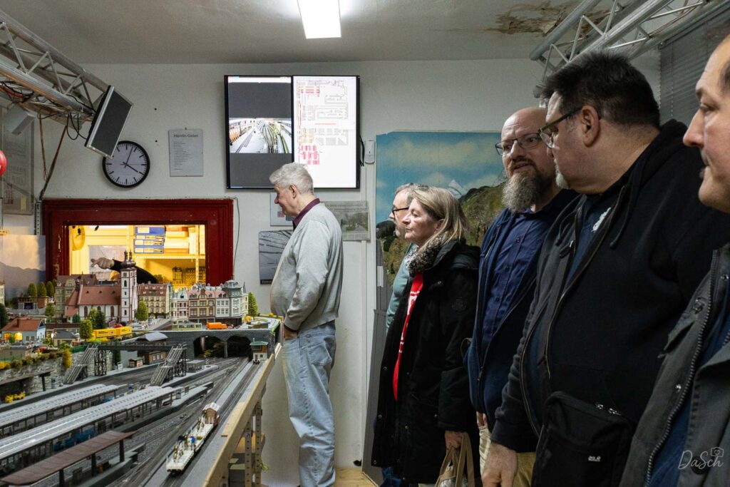 Eine Gruppe erwachsener Besucher betrachtet die Modellbahnanlage mit ihren Gleisen, Gebäuden und der Landschaftsgestaltung.