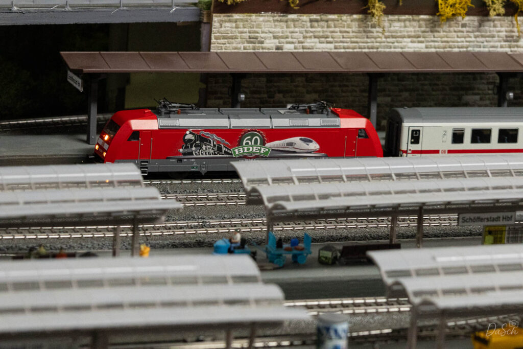 Rote Modelllokomotive mit Jubiläumsbeklebung auf einem Gleis hinter den Bahnsteigdächern des Modellbahnhofs.