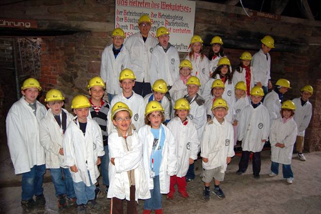 Kinder und Betreuer mit gelben Schutzhelmen und weißen Schutzkitteln bei einer Besichtigung.