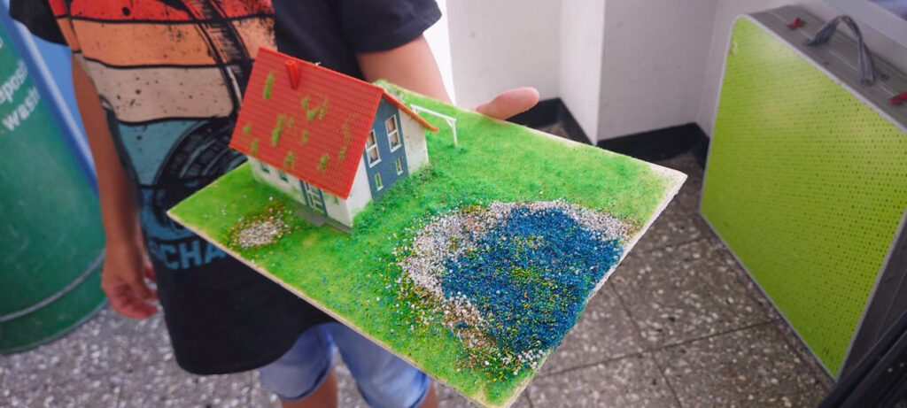 Ein Kind hält eine selbst gestaltete Modelllandschaft mit blauem Haus, rotem Dach, grüner Wiese und blauem Teich.