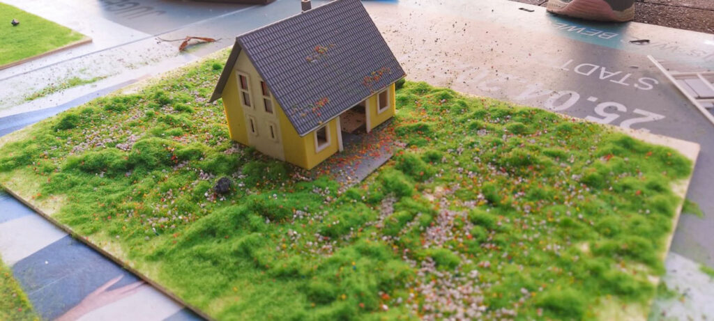 Ein gelbes Modellhaus mit dunklem Dach steht auf einer Grundplatte mit grüner Wiese, kleinen Sträuchern und einem Weg.