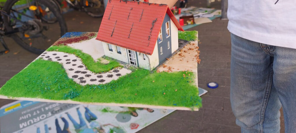 Ein weißes Modellhaus mit rotem Dach steht auf einer begrünten Grundplatte mit geschwungenem Gartenweg und kleinen Steinflächen.