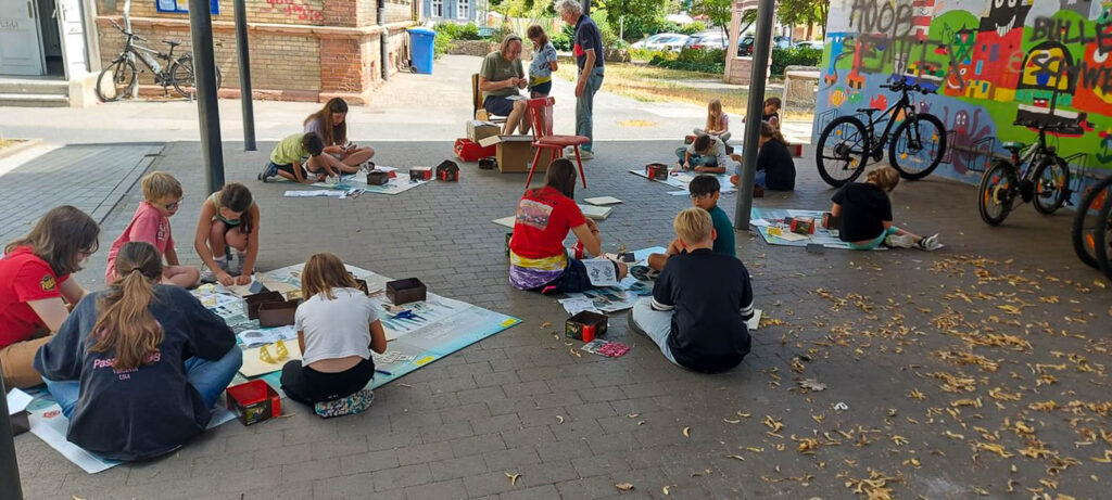 Mehrere Kinder und Betreuer sitzen während der Gernsheimer Ferienspiele im Freien auf dem Boden und arbeiten an ihren Modellbauprojekten.
