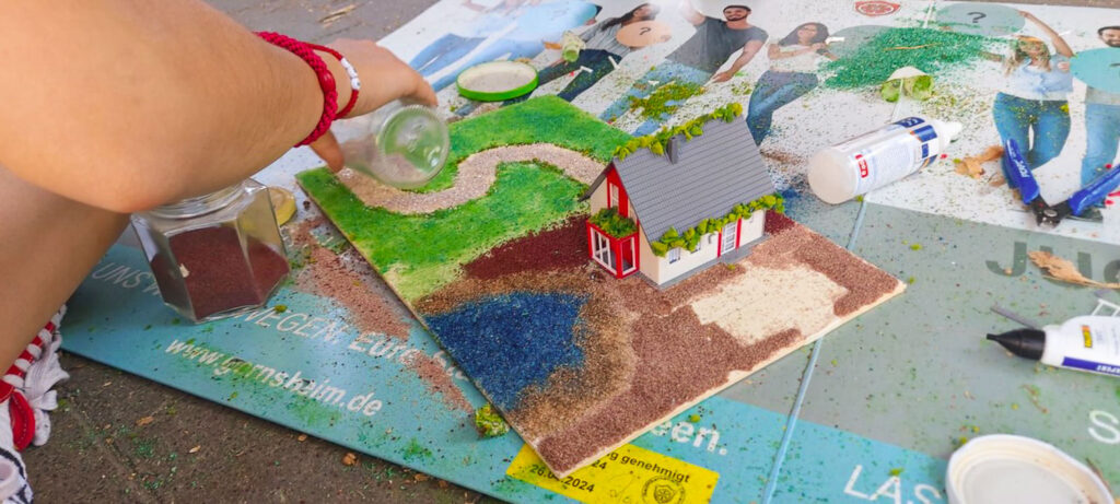 Eine Kinderhand verteilt grünes Streumaterial auf einer Modelllandschaft mit einem kleinen rot-weißen Haus und einem blauen Teich.