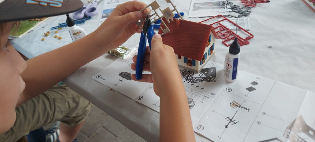 Ein Kind baut bei den Ferienspielen 2026 ein kleines Modellhaus zusammen und bearbeitet die Bauteile mit einer Zange. Auf dem Tisch liegen Bastelmaterial und eine Bauanleitung.