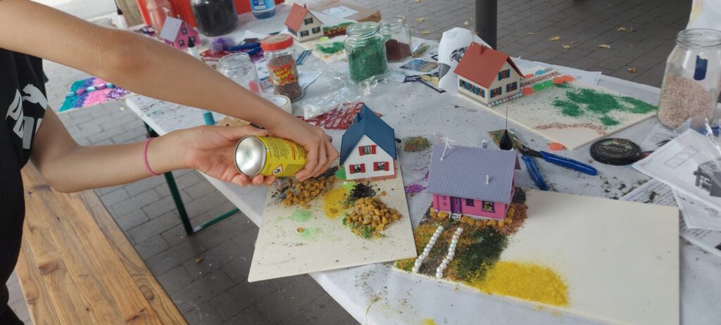 Ein Kind streut aus einer Dose Landschaftsmaterial auf ein selbst gestaltetes Modellgrundstück mit einem kleinen Haus, Garten und Büschen.