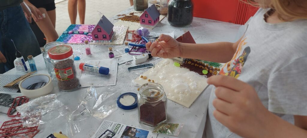 Ein Kind gestaltet bei den Ferienspielen ein Modellgrundstück mit kleinen Dekorationen. Auf dem Basteltisch stehen bunte Modellhäuser, Glitzerstreu, Klebstoff und weitere Bastelmaterialien.