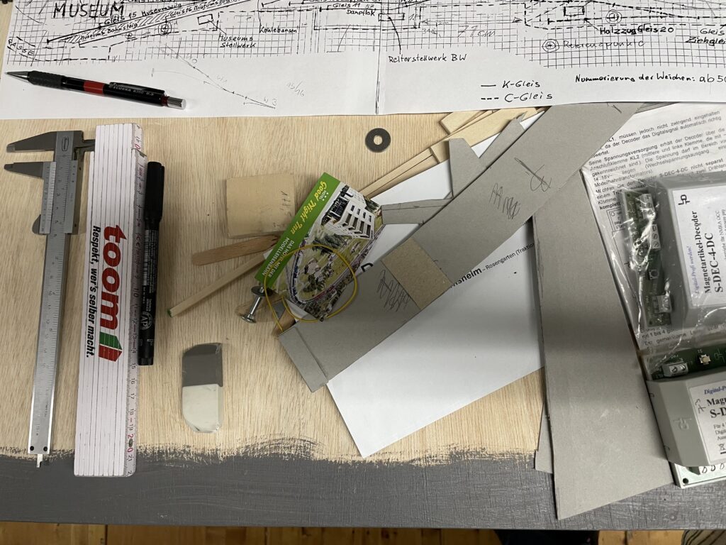 Auf einer Arbeitsfläche liegen Gleispläne für den Bahnhof Rosengarten, Holzleisten, zugeschnittene Platten und weiteres Material für den Bau der Modellbahnanlage.