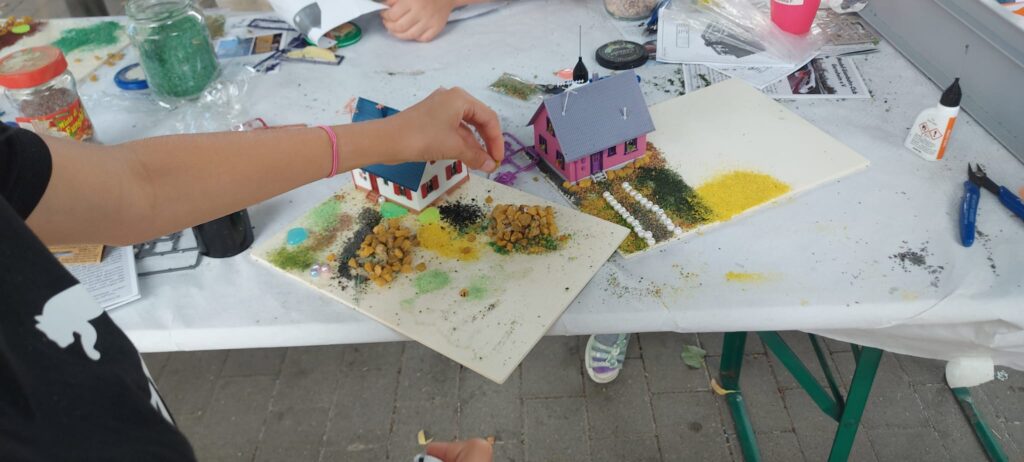 Ein Kind gestaltet bei den Ferienspielen ein kleines Modellgrundstück mit einem Haus, Büschen, Streumaterial und farbigen Landschaftsflächen.